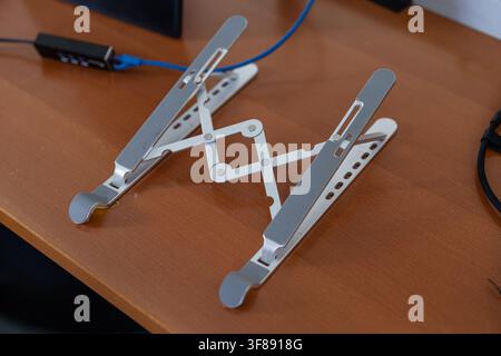 An adjustable, silver aluminum laptop stand set up on a wooden desk surface, featuring anti-slip rubber pads and a foldable scissor mechanism in an Stock Photo