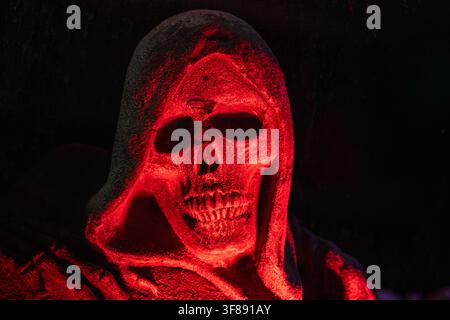 Scary grim reaper skull statue in a hooded cloak illuminated by eerie red light on black background copy space Stock Photo