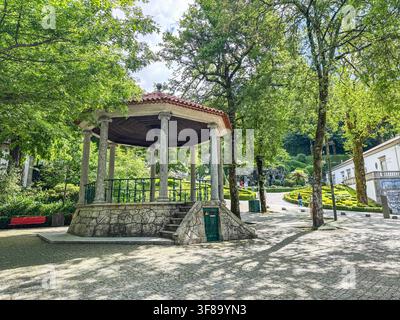 Bom Jesus do monte Pavilion with stone base and red-tiled roof, surrounded by green trees. Sunlight filters through leaves, casting dappled shadows on Stock Photo