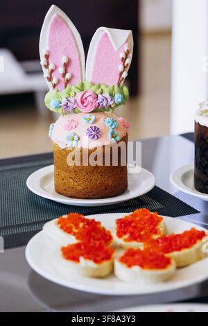 Easter festive table setting with traditional food, cupcakes and red wine at home. Decorated Easter cupcakes with bunny toppers, cheese, candle and re Stock Photo