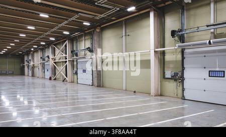 Empty warehouse interior features smooth concrete flooring with white lines. High ceiling has overhead lights and exposed metal beams. Industrial door Stock Photo