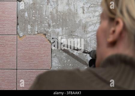 Person using a chisel attachment on a power drill, removing old decorative tiles from a concrete wall during a home renovation or construction project Stock Photo