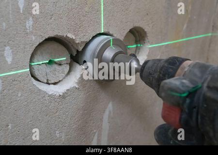 Worker drilling holes in a concrete wall with a powerful rotary hammer and hole saw attachment, using a green laser level for precise positioning duri Stock Photo