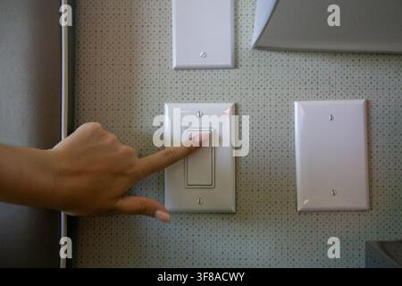 Close-up of a person's hand pressing a standard white decorator-style rocker light switch mounted on a textured interior wall alongside blank electric Stock Photo