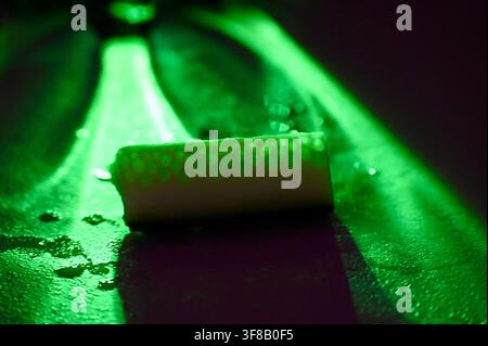 Commercial macro photograph of a vibrant neon green light beam illuminating the finely textured and wet, glistening skin of a fresh cucumber. The focused light creates a stark contrast, highlighting the moisture and intricate patterns on the cucumber's surface against a dark, atmospheric background. Stock Photo