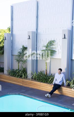 Man sitting on wooden planter bench leaning back, looking right toward turquoise pool, copy space. Outdoor, courtyard, residential, sunlit, shadows, p Stock Photo
