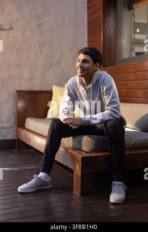 African American man sitting on bench on porch with green pillow wearing gray hoodie, sneakers. Person, patio, deck, lounging, cushions, rustic, casua Stock Photo