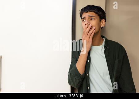 Adult male standing in kitchen by fridge, covering mouth, wearing green shirt, copy space. Refrigerator, door, beige, narrow, appliance, earring, gaze Stock Photo