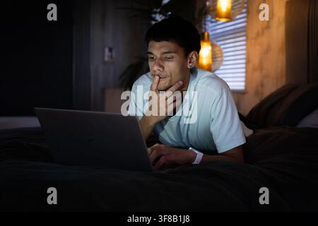 African American man lying on bed, propping on elbows, looking at laptop by pendant lamp. Bedroom, night, ambient, cozy, modern, homeoffice, focus Stock Photo