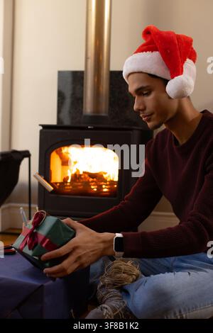 African American man wearing Santa hat sitting by wood stove wrapping gift with twine, copy space. Present, box, cozy, fireplace, hearth, glow, festiv Stock Photo