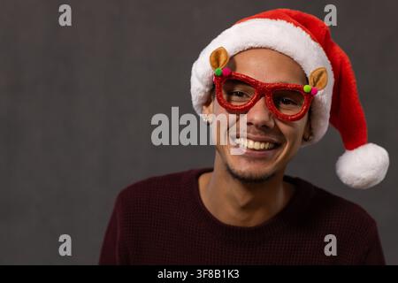 African man smiling wearing red Santa hat reindeer glasses maroon sweater and hoops in studio. Portrait, festive, seasonal, cheerful, accessory, minim Stock Photo