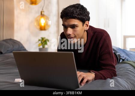 African American man lying on stomach on bed, using laptop, wearing maroon knit sweater and watch. Bedroom, remote, work, calm, focus, cozy, warmth Stock Photo