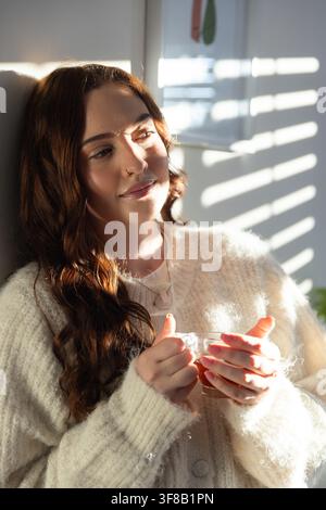 Female sitting in sunlit room holding clear glass mug, wearing fluffy cream sweater, looking down. Woman, cozy, warmth, texture, cup, home, calm Stock Photo