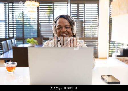 Senior African American woman wearing white headphones, typing on laptop at home with tea. Remotework, sunlit, workspace, shutters, plant, modern, war Stock Photo
