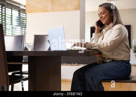African American woman sitting at dining table in home using laptop and phone wearing light shirt. Mature, professional, remote, multitask, productivi Stock Photo