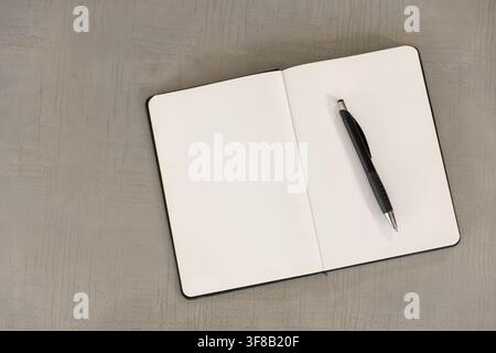 Open notebook lying on textured grey tabletop with blank pages and black pen resting diagonally. Desk, stationery, flatlay, journal, notepad, minimali Stock Photo