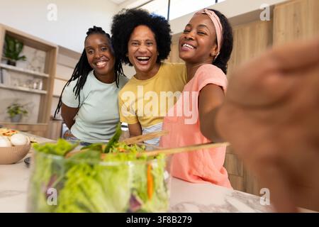 Three friends smiling and posing at kitchen island holding phone, glass salad bowl, wooden tongs. Selfie, candid, lifestyle, pastel, greens, counterto Stock Photo