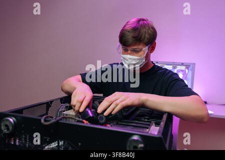 Young man in protective gear assembles desktop PC. Concentrated technician works on hardware upgrade in room with vibrant purple neon lighting Stock Photo