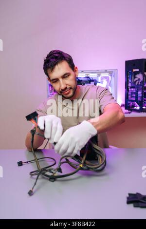 Focused technician in white gloves works on computer hardware. Man assembles custom gaming PC at desk, sorting power supply cables. Purple neon light Stock Photo