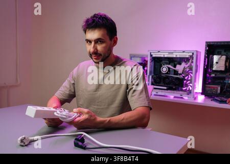 Young man looking at camera while assembling custom desktop PC. He holding power extension cord in hands. Workspace has modern computers with vibrant Stock Photo