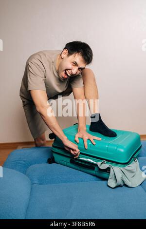 Vertical portrait of fury traveler male struggling to zip overpacked green suitcase for a trip, frowning and making a funny face, experiencing Stock Photo