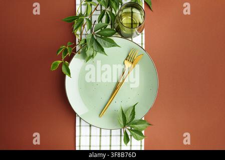 Minimalist studio arrangement features light green speckled plate with golden fork and knife centered on checkered cloth Green vine leaves accent dini Stock Photo