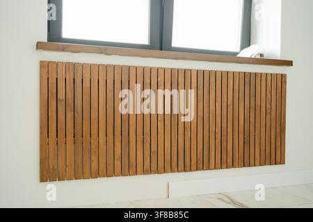 Close-up of vertical wooden slats masking a radiator system under a window. Great visual asset for interior architecture, carpentry design, heating Stock Photo