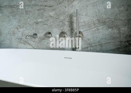 White soaking tub and wall-embedded minimalist faucet controls in a modern stone bathroom interior. Perfect stock photo for relaxation concepts, real Stock Photo