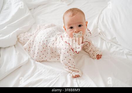 Adorable baby with pacifier looking directly into camera while lying on white bed sheets in a bright room. Perfect image for children's apparel Stock Photo