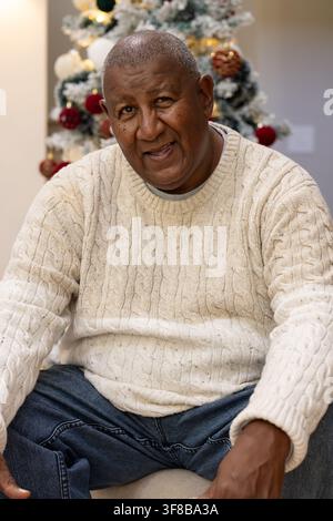 Senior African American man sitting at home, leaning forward in cable-knit sweater by flocked tree. Elder, gentleman, festive, warmth, ornaments, cozy Stock Photo