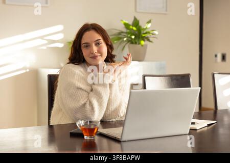 Woman in cream sweater sitting at table in home office, working on laptop with notebook. Female, workspace, dining, sunlit, cozy, focus, minimalist Stock Photo