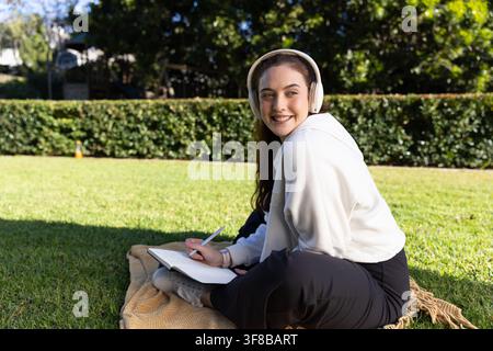 Adult woman sitting on blanket on lawn in hoodie, writing in notebook while listening on headset. Outdoors, headphones, journal, study, park, sunlit, Stock Photo