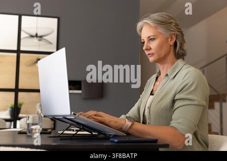 30s woman typing on laptop on angled stand at home desk wearing green shirt, copy space. Computer, workstation, homeoffice, productivity, focus, moder Stock Photo