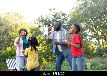 African American family blowing soap bubbles on green lawn in garden holding bubble wands. Yard, outdoor, sunlit, backlit, striped, chair, shrubs Stock Photo