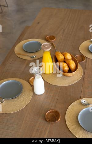 Wooden table is hosting croissant basket, milk carafe and sealed orange juice in dining area. Breakfast, ceramic, plates, surface, natural, light, min Stock Photo