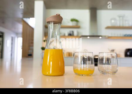 Glass carafe sitting two-thirds full of orange juice on kitchen counter, with two stemless glasses. Beverage, modern, home, surface, shelves, condensa Stock Photo
