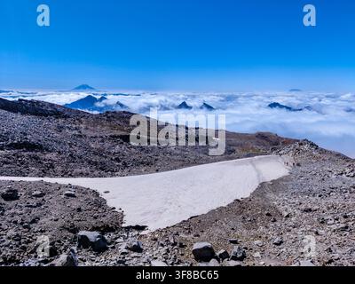 Clear blue sky above rugged mountain peak. Snow patch lines rocky terrain, untouched and pristine. Clouds blanket valley below like a soft white sea Stock Photo