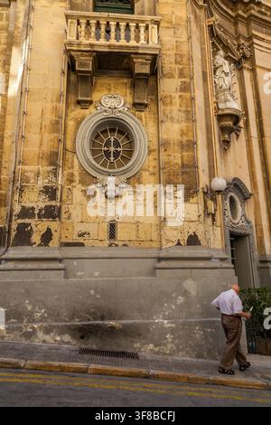A man walks past a weathered baroque facade of Church of St Barbara with an ornate rose window and carved saint statue in Valletta, Malta Stock Photo