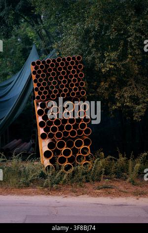 Orange pvc pipes are neatly stacked on wooden pallets, forming a regular pattern of circular openings, with lush green foliage and a tarp visible in t Stock Photo