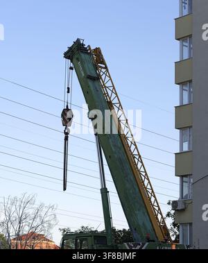 Industrial crane boom and hook extending against a clear blue sky, working on a construction project near a multi story building and power lines Stock Photo