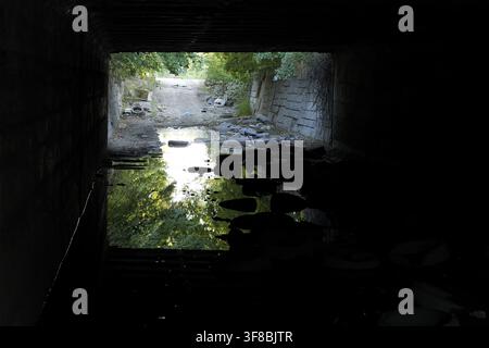 View from inside a dark underpass tunnel showing a small opening with bright light and green trees reflecting on murky water, leading to an unknown pa Stock Photo