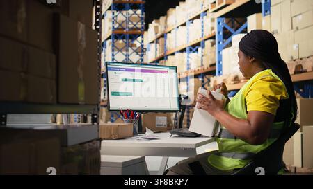 African american woman printing and applying airway bill tags on boxes, wearing hi vis vest in a warehouse near racks. Managing barcode labels with tracking info for order fulfillment. Camera A. Stock Photo