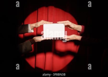 Two female hands present a white box against a red curtain backdrop, symbolizing collaboration in a business presentation setting. Stock Photo