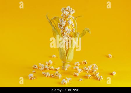 Whole ear of corn stands on a yellow surface with popped popcorn attached and scattered, illustrating popcorn making, snack ingredients and fun food Stock Photo