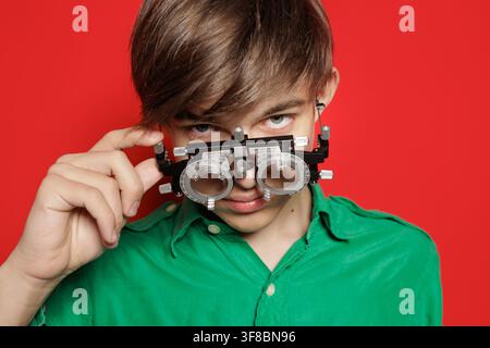 Teen boy in green shirt adjusts trial frame for eye examination against red wall, illustrating vision correction, refractive errors, and optometry Stock Photo
