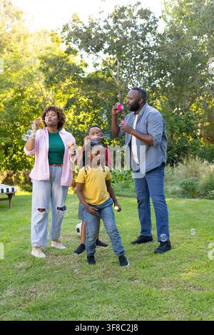 African American family with children blowing bubbles using wands in yard, soccer ball near bench. Outdoor, lawn, garden, sunlit, play, group, candid Stock Photo