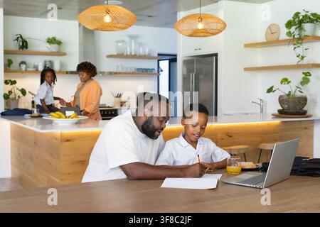African American family studying in modern kitchen, father helping son with laptop and pencil. Homework, computer, table, island, education, backpack, Stock Photo