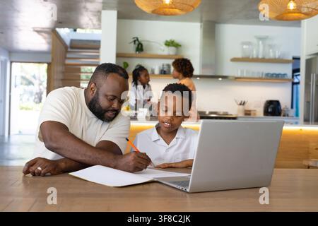 African family father helping youth son in white shirt with notebook at open-plan kitchen laptop. Study, homework, mentoring, learning, daylight, mode Stock Photo