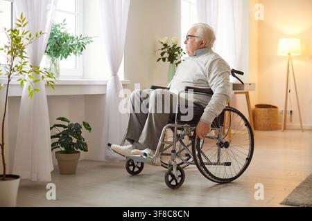 Senior elderly man sitting in wheelchair, looking pensively through window Stock Photo