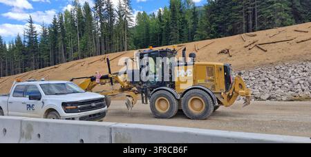 TRANS-CANADA HIGHWAY, CANADA - 10 JULY 2026: A heavy-duty yellow CAT motor grader stands next to a white Ford service pickup truck on a dirt roadbed. Stock Photo
